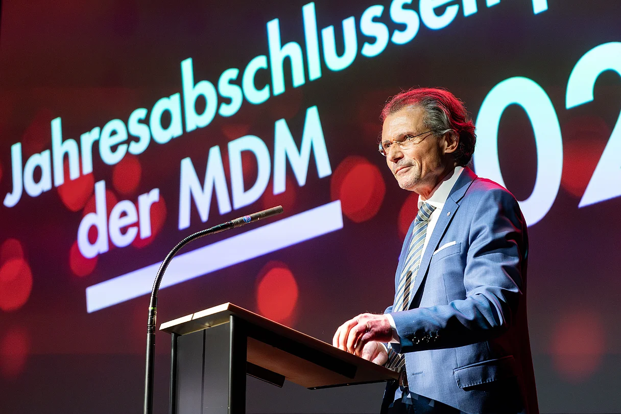 MDM Jahresabschlussempfang 2025 Redner steht an einem Podium auf einer Bühne, im Hintergrund eine Leinwand mit der Aufschrift „Jahresabschlussempfang der MDM 2025“.