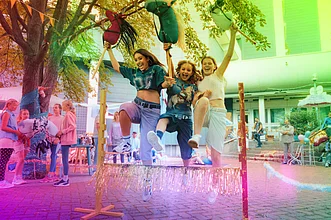 Drei junge Frauen springen lachend über ein Hindernis bei einem Straßenfest und halten bunte Steckenpferde in die Höhe; im Hintergrund stehen weitere Menschen und Dekorationen.