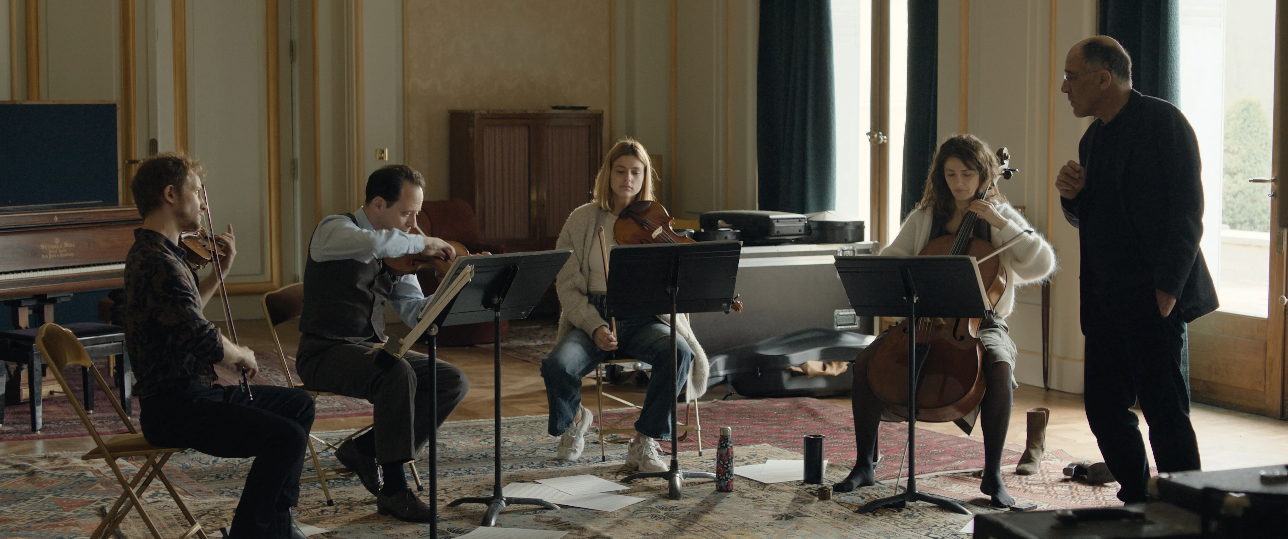 Vier Musikerinnen und Musiker sitzen mit Streichinstrumenten in einem Probenraum, während ein Dirigent oder Dozent ihnen Anweisungen gibt. Vor ihnen stehen Notenpulte, im Hintergrund sind ein Klavier und Instrumentenkoffer zu sehen.