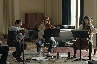 Vier Musikerinnen und Musiker sitzen mit Streichinstrumenten in einem Probenraum, während ein Dirigent oder Dozent ihnen Anweisungen gibt. Vor ihnen stehen Notenpulte, im Hintergrund sind ein Klavier und Instrumentenkoffer zu sehen.