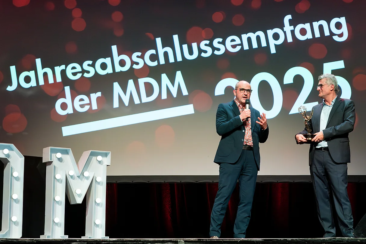 MDM Jahresabschlussempfang 2025 Zwei Personen stehen auf der Bühne vor der Leinwand „Jahresabschlussempfang der MDM 2025“, eine Person spricht ins Mikrofon, die andere hält eine Auszeichnung.