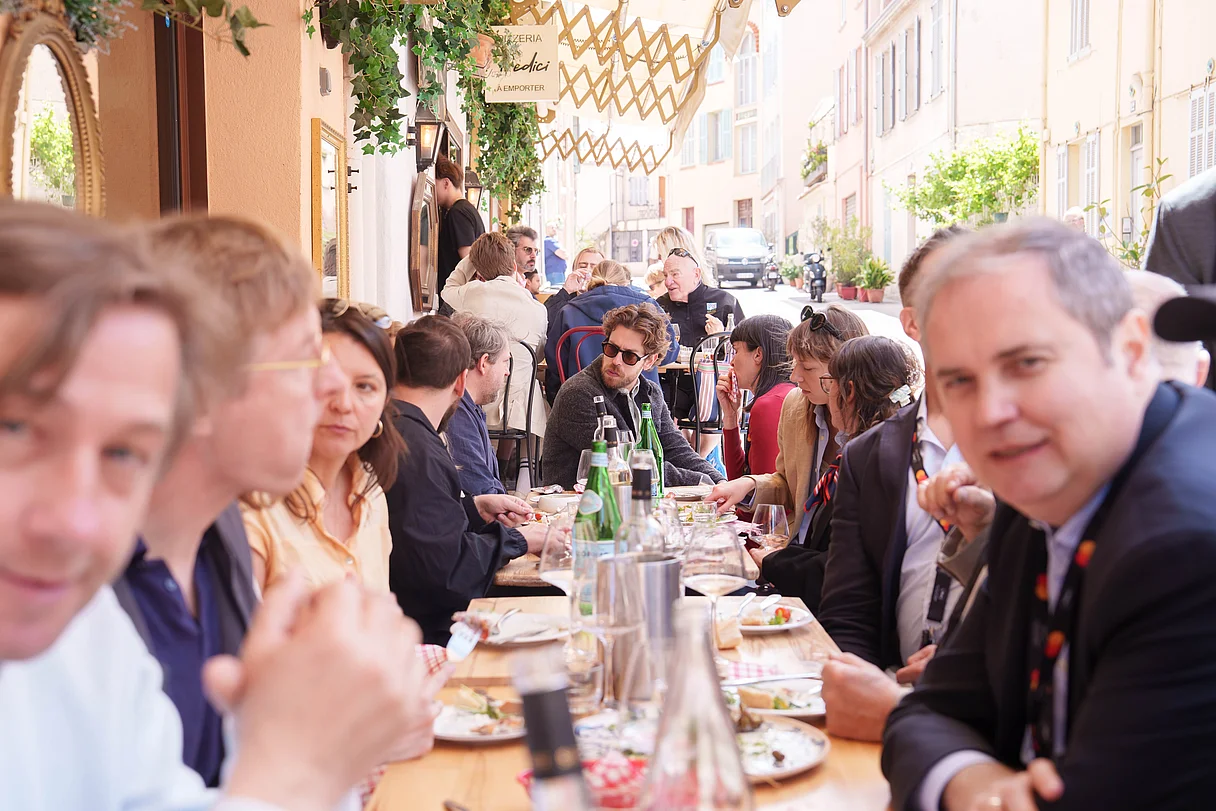 Producers Lunch Cannes 2026 Menschen sitzen an einer langen Tafel vor einem Restaurant in einer schmalen, sonnigen Straße und essen gemeinsam. Im Vordergrund schauen einige Gäste direkt in die Kamera.