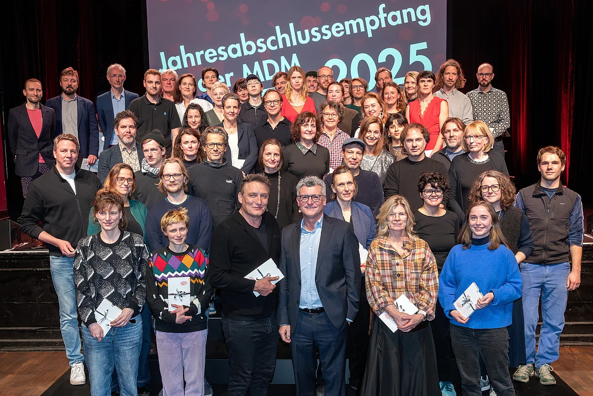 MDM Jahresabschlussempfang 2025 Großes Gruppenfoto mit vielen Teilnehmenden auf einer Bühne, im Hintergrund die Leinwand „Jahresabschlussempfang der MDM 2025“.