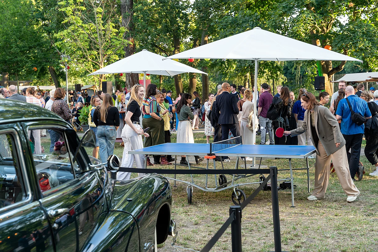 MDM Sommerfest 2026 Menschen stehen und unterhalten sich bei einem Sommerfest im Freien neben einem blauen Tischtennistisch und einem alten grünen Oldtimer unter weißen Sonnenschirmen