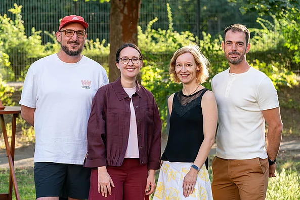 Vier Personen stehen nebeneinander im Freien vor Bäumen, zwei Frauen in der Mitte, links ein Mann mit roter Kappe und weißem T-Shirt, rechts ein Mann mit weißem T-Shirt und brauner Hose