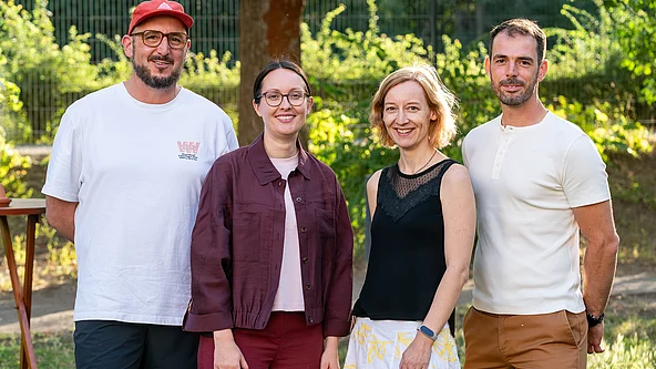 Vier Personen stehen nebeneinander im Freien vor Bäumen, zwei Frauen in der Mitte, links ein Mann mit roter Kappe und weißem T-Shirt, rechts ein Mann mit weißem T-Shirt und brauner Hose