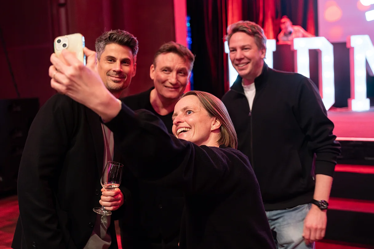 MDM Jahresabschlussempfang 2025 Drei Personen stehen im Hintergrund vor einer Bühne, eine Person im Vordergrund macht ein Selfie.