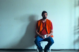 Ein Mann in orangefarbener Warnweste sitzt mit einer Tasse in der Hand auf einem Stuhl vor einer weißen Wand. Die minimalistische Szene wirkt ruhig und nachdenklich.