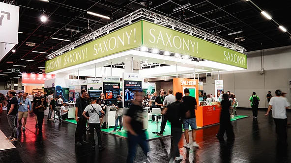 Messestand mit grünem Banner und der Aufschrift »SAXONY!« auf einer Messe mit vielen Besucher*innen