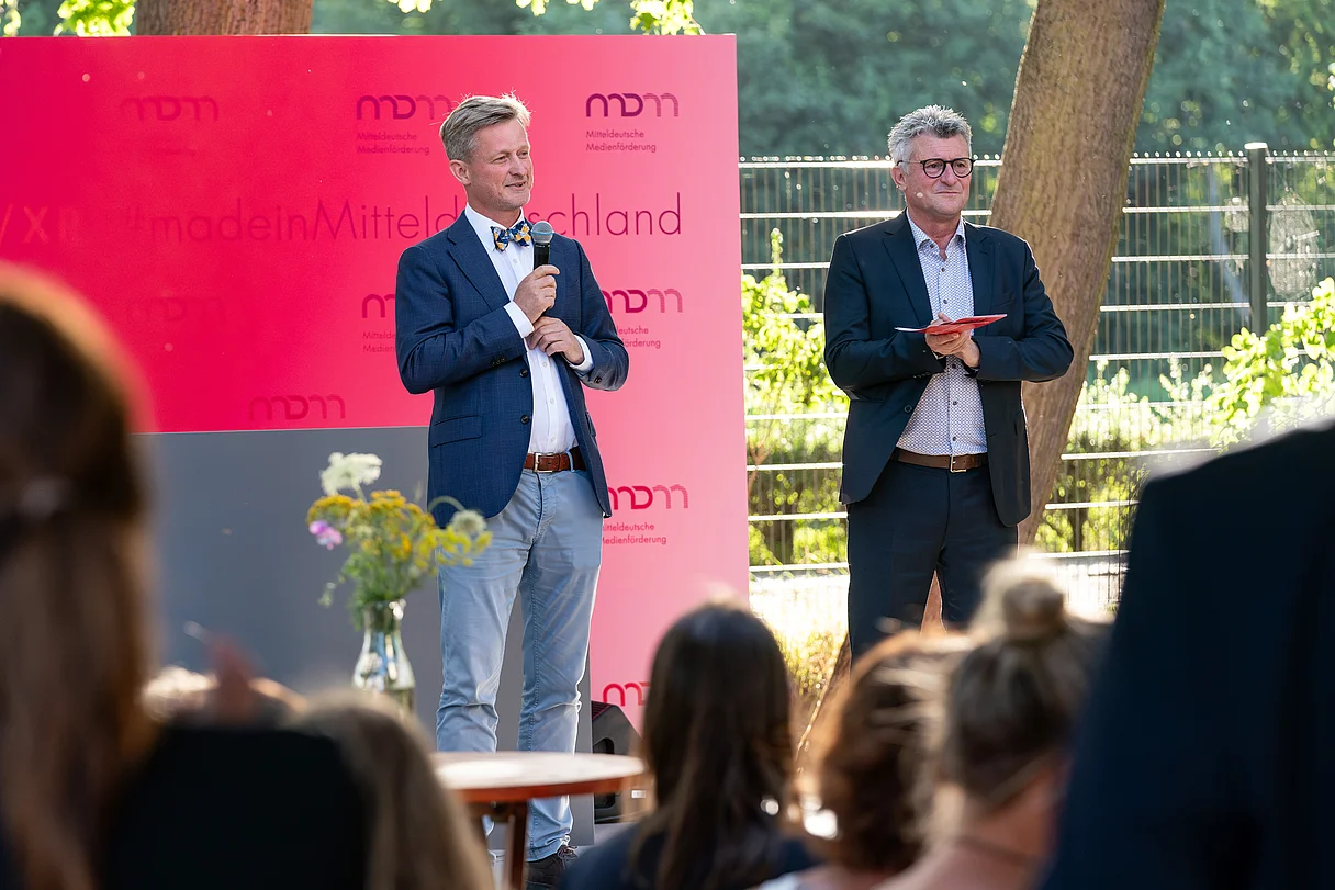 MDM Sommerfest 2026 Zwei Männer in Anzügen stehen vor einem roten Hintergrund mit Schrift und sprechen vor einem Publikum im Freien