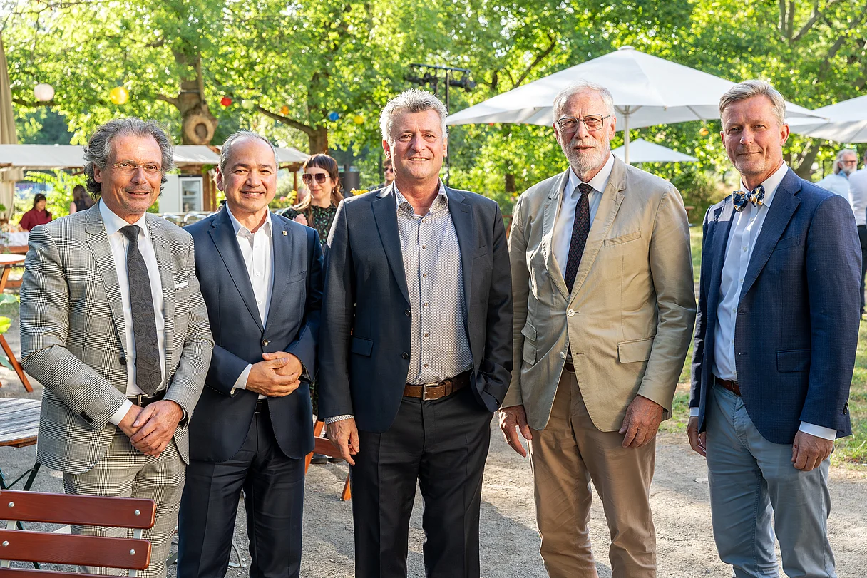 MDM Sommerfest 2026 Fünf Männer in Anzügen stehen im Freien vor Bäumen und Sonnenschirmen und blicken in die Kamera