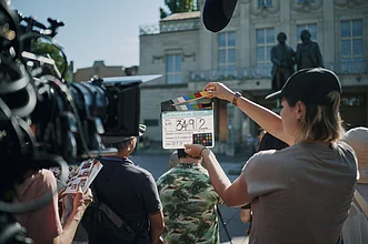 Filmteam bei Dreharbeiten im Freien: Eine Person hält eine Filmklappe vor einer Kamera, im Hintergrund ein Gebäude und mehrere Crewmitglieder.