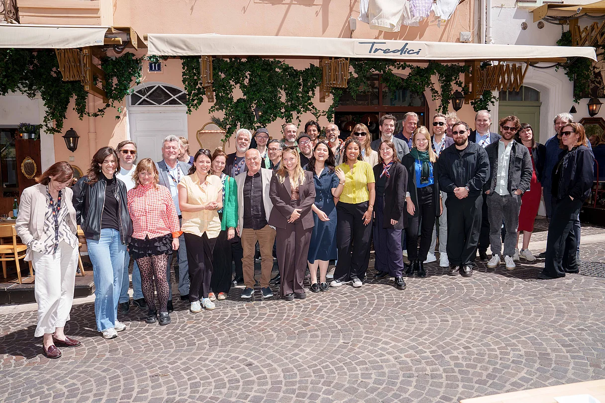 Producers Lunch Cannes 2026 Große Gruppe von Menschen posiert vor einem Restaurant mit Sonnendach in einer mediterranen Altstadtstraße. Die Personen stehen dicht beieinander und blicken lächelnd in die Kamera.