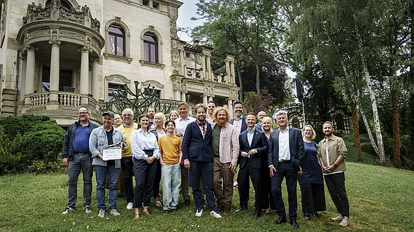 Gruppe von Menschen steht auf einer Wiese vor einem historischen Gebäude mit Säulen und Verzierungen
