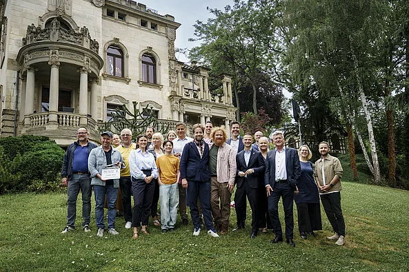 Gruppe von Menschen steht auf einer Wiese vor einem historischen Gebäude mit Säulen und Verzierungen