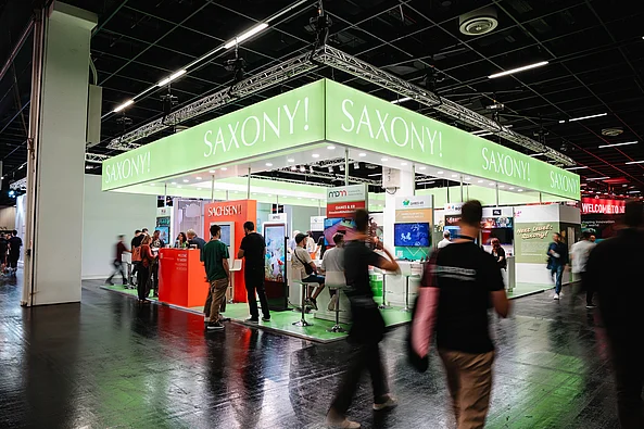 Messestand mit grünem Banner und weißer Aufschrift »SAXONY!« auf einer Messe mit mehreren Personen