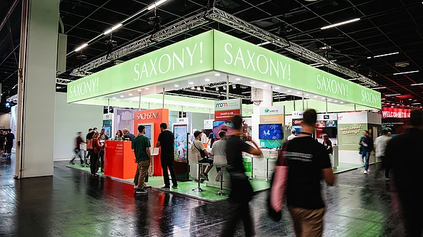 Messestand mit grünem Banner und weißer Aufschrift »SAXONY!« auf einer Messe mit mehreren Personen