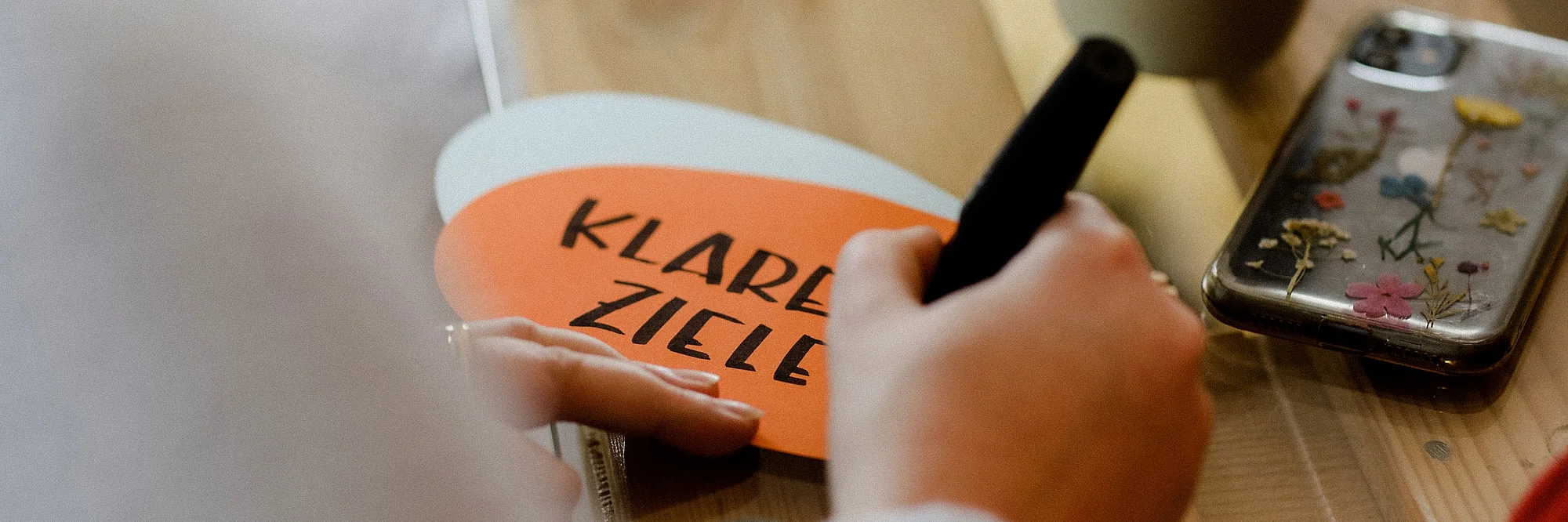 Hand schreibt mit Filzstift auf eine orange Karte mit der Aufschrift „Klare Ziele“, auf einem Tisch mit Smartphone und Kaffeebecher. | Urheberrecht: Anne Schwerin