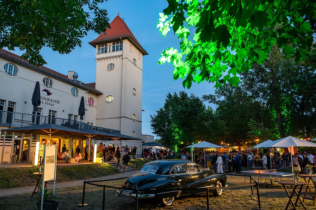 MDM Sommerfest 2026 Menschenmenge bei einem Sommerfest vor einem historischen Gebäude mit Turm und einem schwarz-grünem Oldtimer-Pkw im Vordergrund
