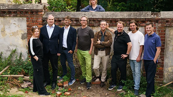 Gruppe von acht Personen steht vor einer alten Mauer mit offenem Tor und einer weiteren Person, die auf der Mauer lehnt
